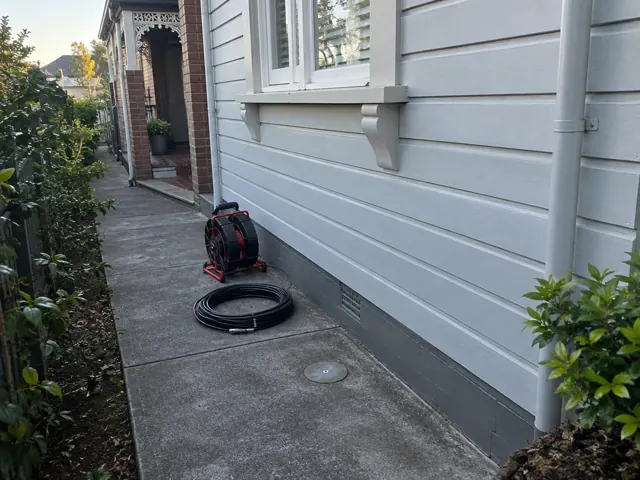 Capped drain inspection opening beside a coiled camera reel on a concrete path by a weatherboard house in Botany
