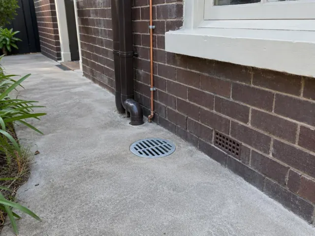Outdoor brick wall and concrete path with a round drain grate and wall vent in Botany