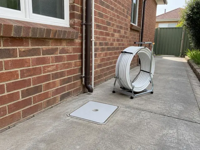 Concrete driveway beside a brick house with a capped inspection cover and pipe reel on a frame in Mascot