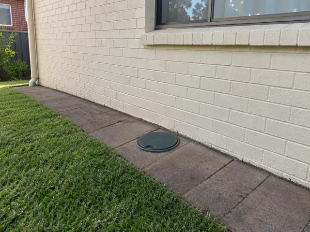 Capped sewer inspection opening in a paved side path beside a cream brick house in Eastlakes